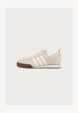 Baskets Adidas beiges à bandes blanches, détail de logo doré, dessus en suède beige, lacets blancs et semelle en caoutchouc marron sur fond uni.
