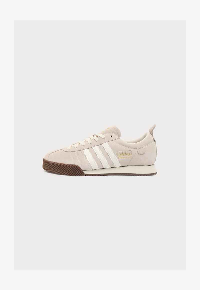 Μπεζ αθλητικό Adidas με λευκές ρίγες, χρυσό λογότυπο, επάνω μέρος από μπεζ σουέτ, λευκά κορδόνια και καφέ λαστιχένια σόλα σε απλό φόντο.