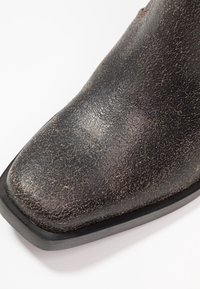 Bota de cuero negro con una superficie texturizada, punta cuadrada y suela baja y plana. Presenta un leve brillo y un acabado resistente.