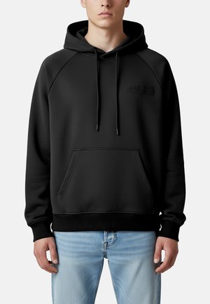 Hombre con sudadera negra con capucha y bolsillo frontal con el logo "4US", combinado con jeans azul claro, de pie frente a un fondo blanco.