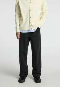 Light yellow knit cardigan with button closure, two front pockets, worn over a light blue striped shirt and black loose-fit jeans. Black shoes.