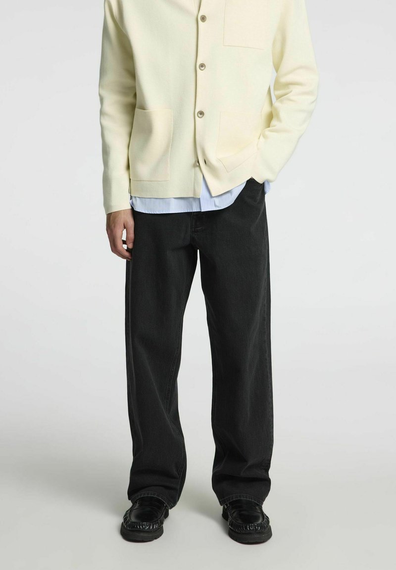 Light yellow knit cardigan with button closure, two front pockets, worn over a light blue striped shirt and black loose-fit jeans. Black shoes.