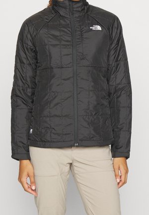 Henkilö, jolla on yllään musta tikattu North Face -takki vetskarilla ja beiget housut yksiväristä vaaleaa taustaa vasten.
