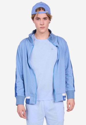 Giovane uomo con capelli castano chiaro indossa un berretto azzurro girato all'indietro, una giacca azzurra con cerniera, una maglietta e pantaloni abbinati, rivolto in avanti.