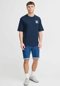 Jūras zila īslais T-krekls ar logo uz priekšpuses, savienots ar rullētām džinsu šortiem. Modelis valkā baltas zeķes un pelēkas sporta kurpes.