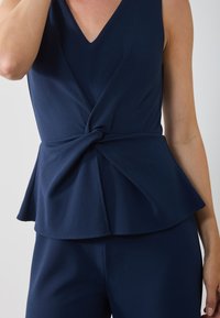 Top peplum bleu marine en tissu doux, doté d'un décolleté en V et d'un détail torsadé à la taille. Texture lisse avec un ourlet fluide.