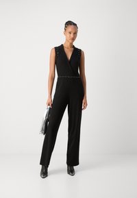 Jumpsuit noir sans manches avec un col, des accents d'épaules cloutés et une taille ceinturée. Associé à des bottines noires à talons et une pochette argentée.