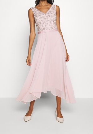 Robe sans manches rose pâle à col en V avec corsage floral brodé, taille cintrée, ourlet asymétrique fluide, portée avec des talons à bout pointu.