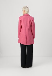 Roze wollen mixjas met een gestructureerd ontwerp, brede revers en een enkele split aan de achterkant. Gecombineerd met zwarte wijde broek.