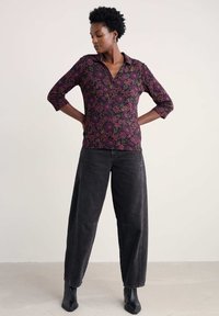 Schwarz geblümtes Hemd mit Kragen und Dreiviertelärmeln, verziert mit lila und grünen Mustern, kombiniert mit locker sitzenden dunklen Jeans und Stiefeletten.
