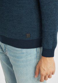 Marineblauer Strickpullover mit strukturiertem Muster, gerippten Bündchen und Saum. Verfügt über ein kleines Logotag nahe dem Saum. Kombiniert mit hellblauen Denim-Jeans.