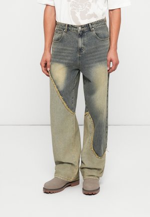 Jeans de pierna ancha desgastados en mezclilla azul y beige. Presenta un diseño de patchwork y acentos deshilachados. Se llevan con botas de tobillo grises.
