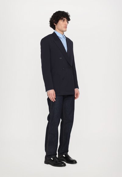 Blazer double-breasted bleu marine avec revers crantés, associé à une chemise bleu clair, un jean en denim foncé et des chaussures en cuir noires.