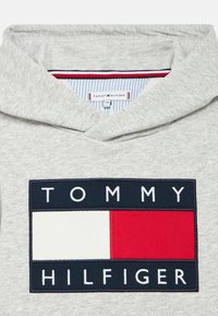 Sweat à capuche gris avec un grand patch logo. Le patch présente des couleurs rouge, blanc et bleu avec "TOMMY" au-dessus et "HILFIGER" en dessous en lettres blanches.