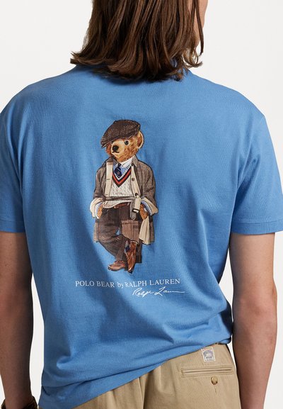 T-shirt din bumbac albastru, cu un grafic ce prezintă un urs îmbrăcat într-un palton și o șapcă, având inscripția "POLO BEAR by RALPH LAUREN" imprimată dedesubt.