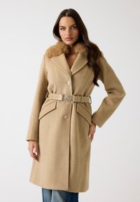 Cappotto in misto lana beige con colletto in pelliccia, bottoni doppi e vita con cintura. Presenta tasche a patta e una silhouette su misura.