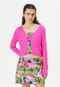 Cardigan corto rosa con maniche a palloncino e legatura anteriore, abbinato a una gonna a fantasia floreale dai colori vivaci. Tessuto morbido e design aderente.
