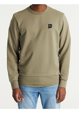Hombre con sudadera de manga larga verde oliva con parche negro con el logo en el pecho y vaqueros azules claros, de pie frente a un fondo liso.