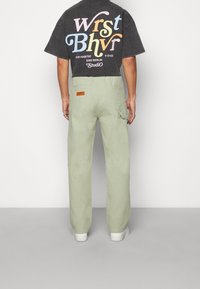 Grå grafisk t-shirt med flerfärgad text på baksidan, tillsammans med ljusgröna byxor med bakficka och logomärke. Vita skor är synliga.