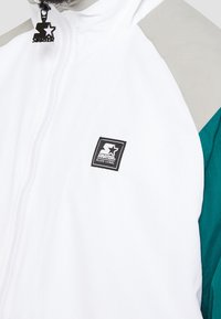 Biała kurtka z akcentami w kolorze morskim i szarym; posiada zamek błyskawiczny i naszywkę z logo. Materiał wydaje się być lekki o gładkiej teksturze.