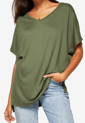 Donna che indossa una maglietta verde oliva a maniche corte con scollo a V e jeans azzurri, con una mano in tasca e una collana d'oro visibile.