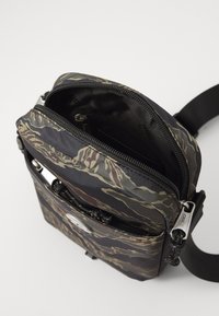 Piccola borsa a tracolla con motivo mimetico, scomparto principale parzialmente aperto e tasca frontale con zip, cinghia nera regolabile visibile.