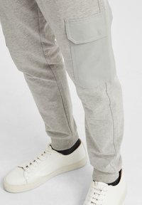 Grå joggebukser med en sidelomme i lysegrå stoff, tapered design og en glatt tekstur, sammen med hvite sneakers.