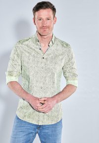 Hellgrünes Hemd mit Blattmuster, lange Ärmel bis zu den Ellbogen hochgekrempelt, getragen mit blauen Jeans, Hände vor sich gefaltet.