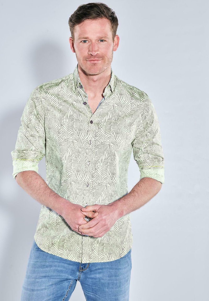 Hellgrünes Hemd mit Blattmuster, lange Ärmel bis zu den Ellbogen hochgekrempelt, getragen mit blauen Jeans, Hände vor sich gefaltet.