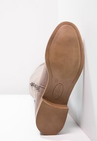 Beżowy zamszowy botek na kostkę z gładką podeszwą, z zaokrąglonym noskiem i prostym przeszyciem. Podeszwa ma teksturowany bieżnik i detal z logo.