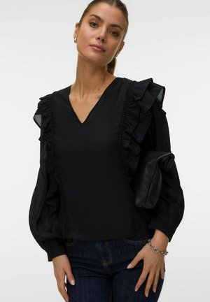 Kobieta z włosami związanymi do tyłu, ubrana w czarną bluzkę z falbankami na rękawach oraz ciemne dżinsy, trzymająca złożoną czarną kopertówkę pod pachą.