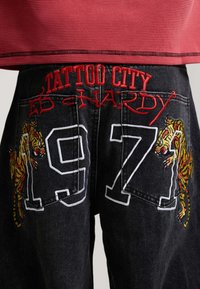 Pantaloni in denim nero con ricamo rosso "TATTOO CITY ED HARDY" sopra le tasche, numeri bianchi "1971" e toppe gialle con tigre su ciascun lato.