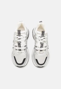 Białe i szare sneakersy z siateczkową cholewką, grubą podeszwą, odblaskowymi akcentami oraz czarnymi detalami na sznurowadłach i boku.