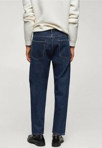 Jeans blu scuro con taglio dritto, dotati di due tasche posteriori e dettagli di cucitura sottili, indossati con un maglione di colore chiaro e scarpe nere.