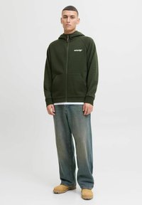 Haut à capuche zippé vert foncé avec des accents côtelés, poches avant, logo sur la poitrine ; associé à un jean bleu délavé et ample et des bottes beiges.