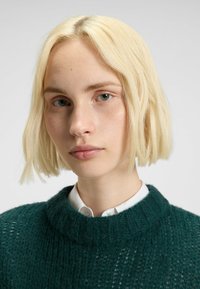 Maglione verde lavorato a maglia con un ampio collo a coste, indossato sopra una camicia bianca con colletto. I capelli biondi, corti e spettinati incorniciano il viso.