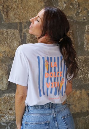 Frau mit braunen Haaren im Pferdeschwanz, die ein weißes T-Shirt mit blauen Streifen, orangefarbener Aufschrift "LA DOLCE VITA" und Eiscreme-Kegel-Grafiken trägt, steht an einer Steinmauer.
