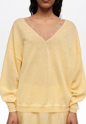 Mujer con suéter de cuello en V amarillo pálido con ribete de encaje beige en el escote, mangas largas sueltas y falda o pantalones a juego.