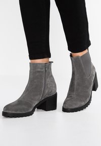 Botas de tobillo de ante gris con cremallera lateral, acentos elásticos y un tacón grueso negro. Suela texturizada que proporciona tracción para mayor estabilidad.