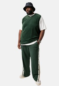 Groene velours mouwloze top over een wit t-shirt, gecombineerd met groene broeken met witte zijstrepen en knopenaccenten, en een donkere buckethat.