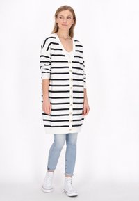 Zwart-wit gestreepte cardigan met een v-hals, lange mouwen en gouden knopen, gecombineerd met lichtblauwe jeans en witte sneakers.