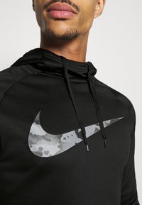 Plano detalle de una persona que lleva una sudadera con capucha negra con un swoosh de Nike de patrón de camuflaje gris y cordones negros.