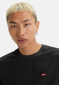 Ung person med kort, afbleget hår, iført guld hoop-øreringe og en sort Levi's skjorte med et lille rødt logo på brystet.