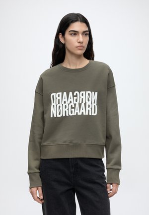Kvinde med langt mørkt hår iført olivengrøn sweatshirt med hvidt spejlet "NØRGAARD" tekst og sorte bukser, stående mod en ensfarvet baggrund.