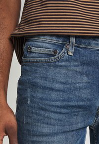 Persona que lleva vaqueros de mezclilla azules y una camisa a rayas marrón y negra, con el brazo izquierdo y la mano visibles.