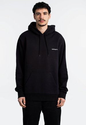 Sudadera negra de algodón con un bolsillo frontal, capucha con cordones y pequeño logo blanco en el pecho. Modelo lleva pantalones negros.