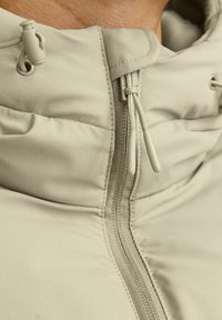 Veste beige clair avec un col haut, dotée d'une fermeture éclair et de cordons de serrage. Le tissu a une texture subtile avec un motif en grille.