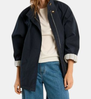 Vrouw die een marineblauwe jas met ritssluiting draagt met opgerolde gestreepte manchetten, een wit overhemd en blauwe spijkerbroek tegen een effen achtergrond.