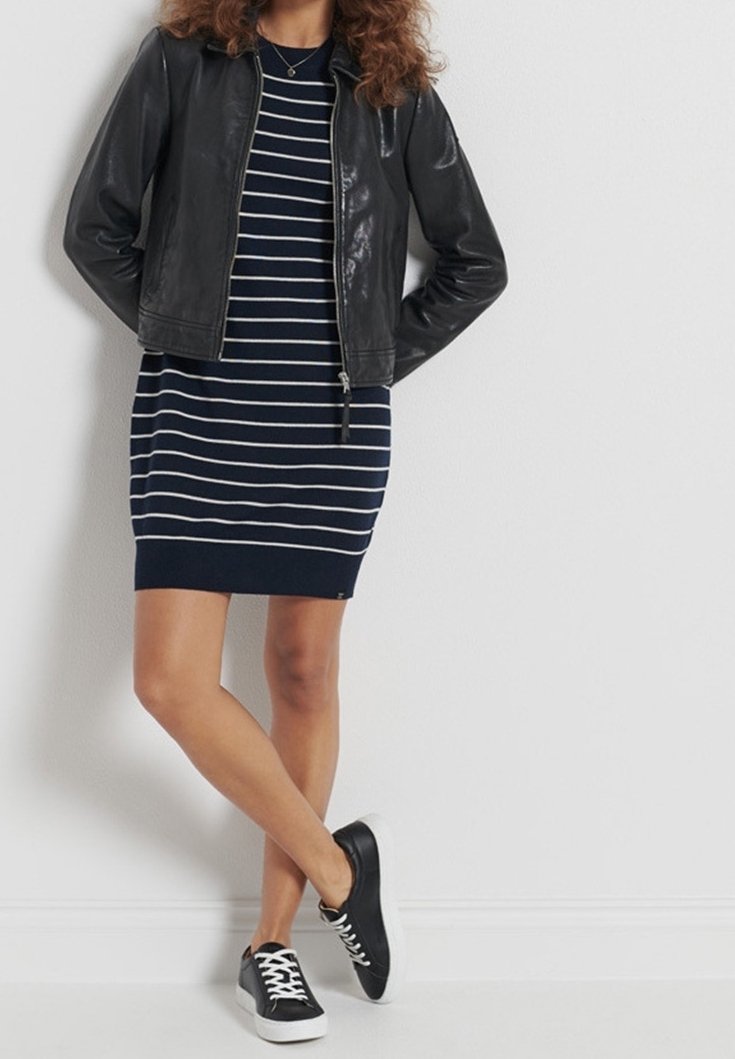 Femme portant une veste en cuir noire sur une robe rayée bleu marine et blanche, associée à des baskets noires à lacets blancs, debout jambes croisées.