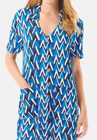 Vestido de manga corta en patrón zigzag en azul, blanco y negro, con escote en V, botonera frontal y dos bolsillos delanteros. Tela suave.